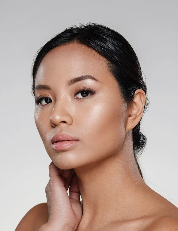 Close-up beauty portrait of a woman with a light-medium skin tone and sleek dark hair, hand resting at her neck, soft grey studio background