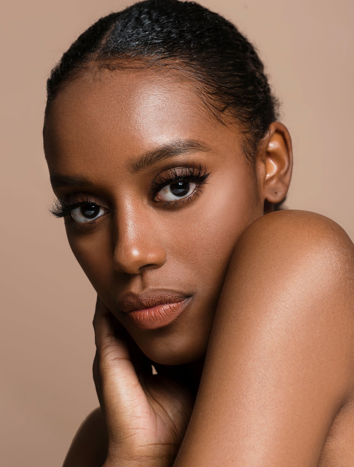 Close-up beauty portrait of a woman with rich, deep skin tone resting her hand against her cheek, warm neutral studio background